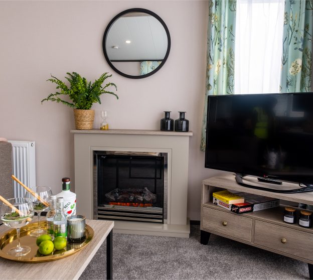 Inviting static caravan interior with tropical plants adorning a fireplace, drinks placed on a coffee table, and a large TV in the corner.