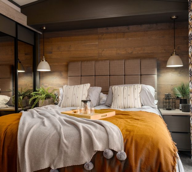 Boho style bed in a static caravan with yellow blanket and breakfast tray on top, flanked by mirrored wardrobe and window.
