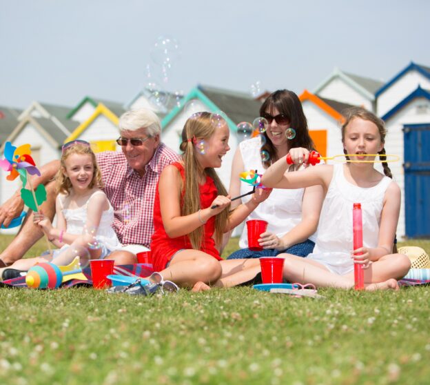 Family sitting on grass in front of beach huts.