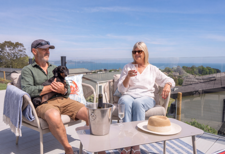 Couple sat on lodge decking with sea view having a glass of prosecco with their dog
