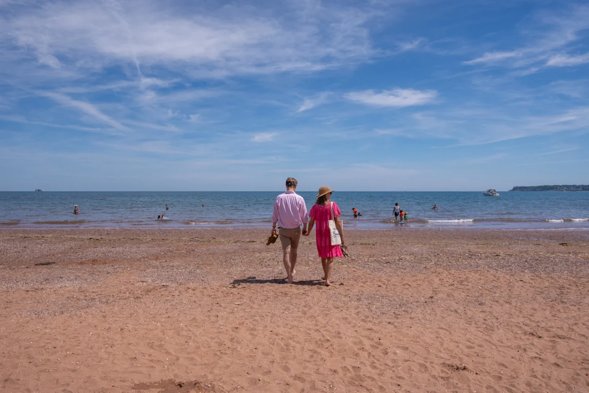 Older couple holding hand walking barefoot on beach at Goodrington Sands.