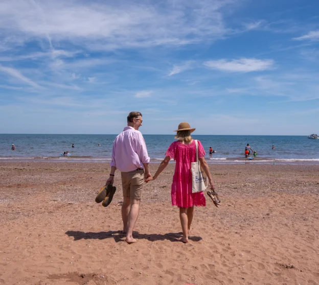 Older couple holding hand walking barefoot on beach at Goodrington Sands.
